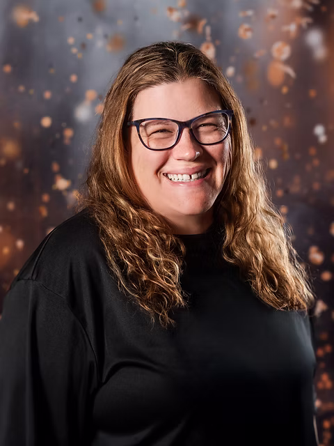 Heather, dance instructor wearing glasses, with long wavy hair and a joyful smile against a sparkling bokeh background