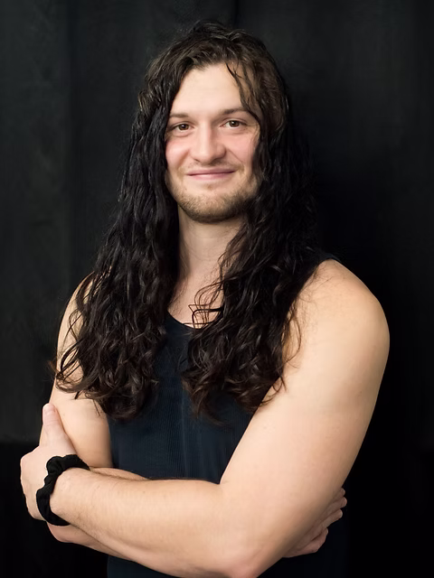 Myrni, a male instructor with long curly dark hair, arms crossed and smiling confidently against a dark studio background