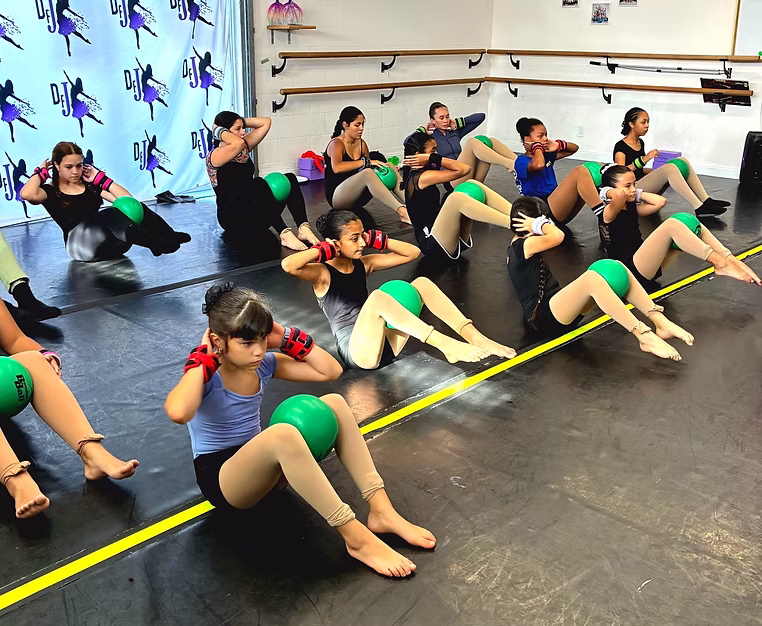 Dancers lie on mats performing abdominal exercises with green stability balls, practicing strength and control together in the studio.