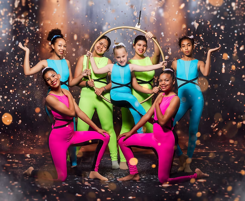 Seven dancers in bright neon unitards pose with an aerial hoop, arranged in energetic standing and seated positions as golden sparkles create a lively, acrobatic atmosphere.