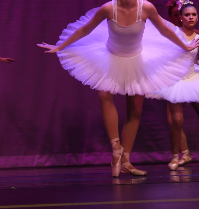 A ballet dancer in a white classical tutu performs on pointe under stage lighting, with additional dancers in similar costumes visible behind her against a purple backdrop.