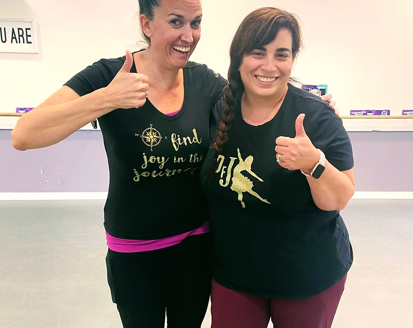 Two smiling adults stand together in a dance studio, giving enthusiastic thumbs-up gestures while wearing matching black shirts, creating a friendly and encouraging atmosphere.