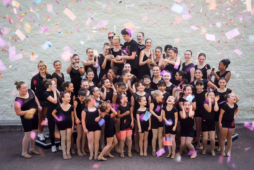 A large group of young dancers in black uniforms celebrates together outdoors, smiling and laughing as colorful confetti falls around them.