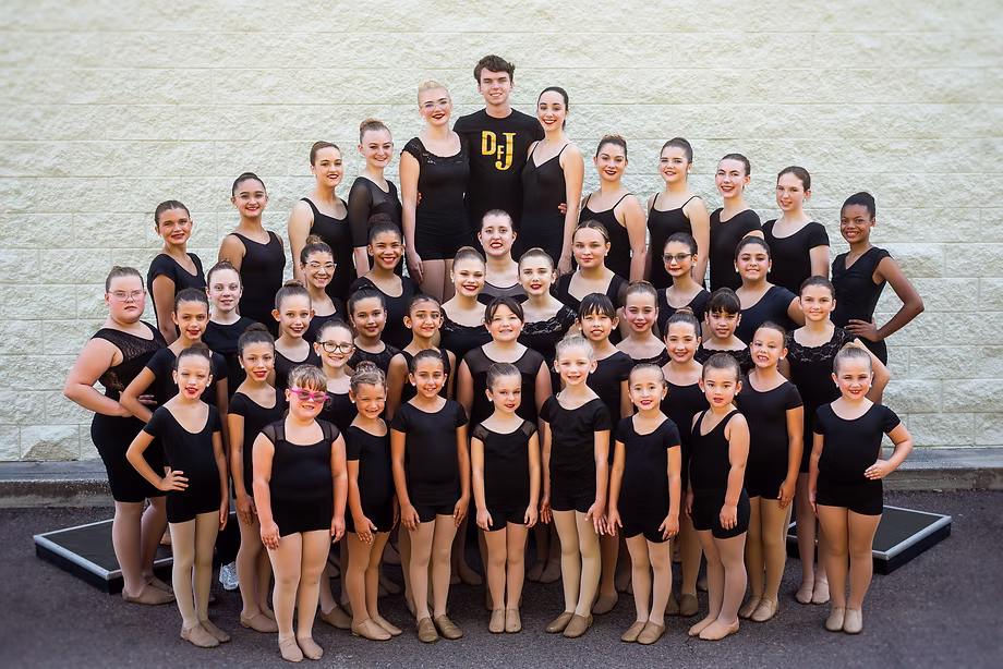 A large group of dancers in black class attire poses in organized rows outdoors, smiling confidently for a formal team photo.