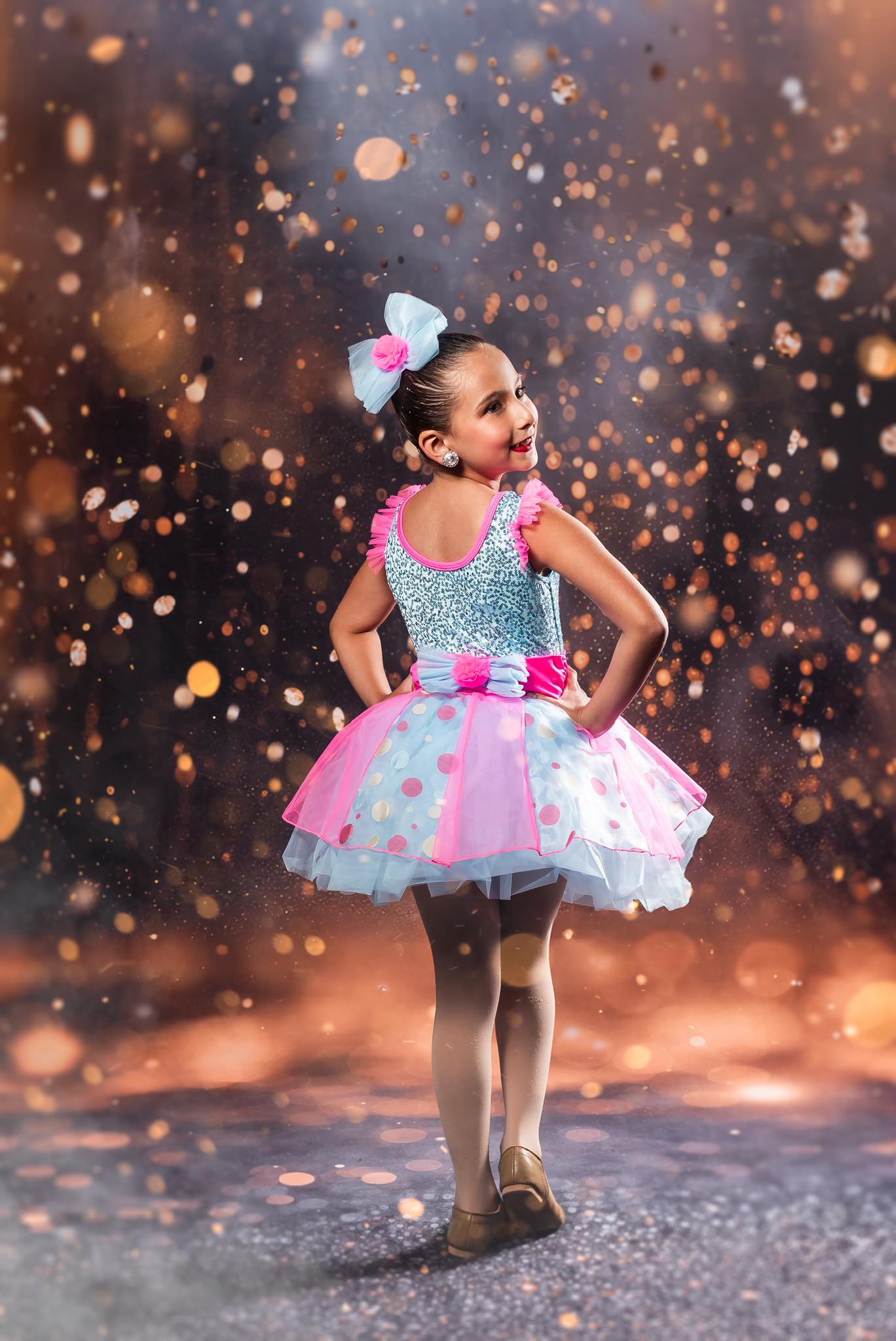 Young girl wearing a sparkly blue and pink tutu costume poses confidently with hands on hips against a warm, glowing background with glitter-like bokeh lights.