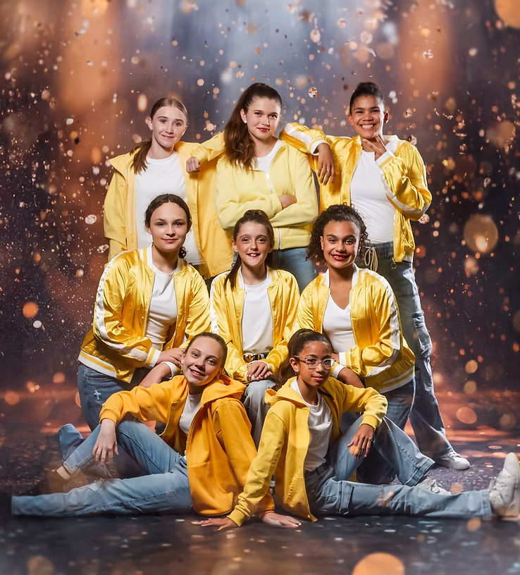 Eight dancers in yellow jackets, white tops, and jeans pose confidently onstage, arranged in standing and seated positions with warm golden lights and sparkles creating an energetic, upbeat atmosphere.