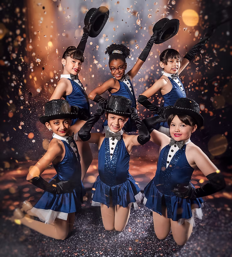 Six young dancers in sparkly blue jazz costumes and black top hats pose confidently onstage, smiling brightly as golden sparkles and warm lighting create an energetic, celebratory atmosphere.