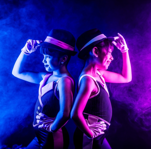 Two dancers in pinstripe vests, gloves, and fedoras stand back-to-back, tipping hats under dramatic blue and pink stage lights.