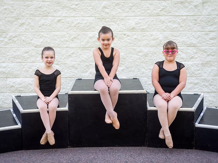 Three young dancers in black leotards sit on outdoor stage blocks, smiling as they pose together for their Joy Bundles team photo.