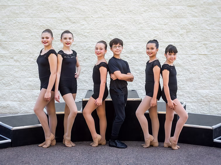 Six dancers in black uniforms pose outdoors, standing and sitting in a line with confident expressions for their Joy Fire team photo.