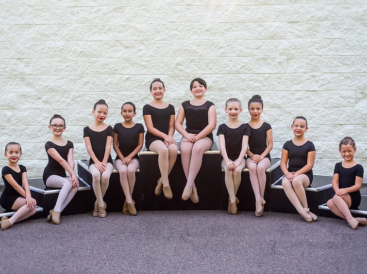 A group of young dancers in black class attire sits in a row outdoors, smiling brightly for their Joy Ignite team picture.