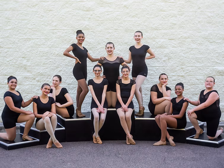 Dancers in black uniforms pose outdoors in seated and standing positions, smiling confidently together for their Joy Intensity team photo.