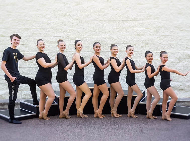 The Joy Lamplighters team poses in a line outdoors wearing black leotards, arranged beside their coach for a clean, unified group photo.