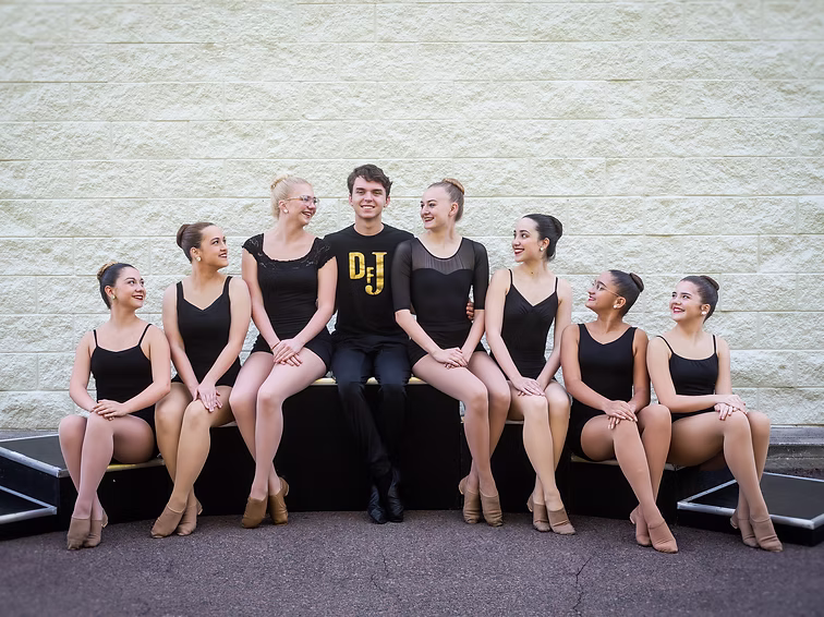 A row of dancers in black attire sits outdoors beside their coach, smiling warmly for the Joy Radiance team portrait.