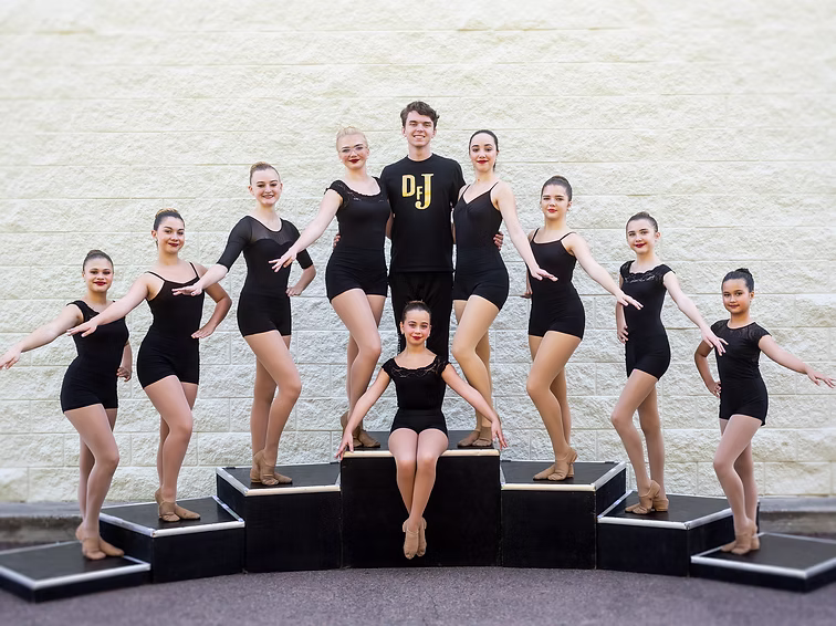 Dancers in black leotards pose on raised blocks outdoors, arranged in dynamic levels for the Joy Soleil team picture.