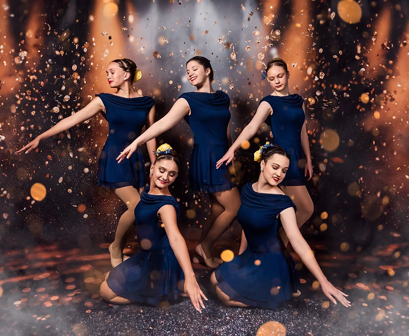 Five dancers in elegant navy dresses perform a soft, lyrical pose onstage, extending their arms gracefully as warm golden lights and sparkles create a dreamy, expressive atmosphere.