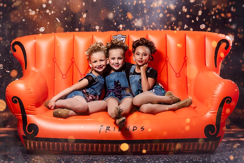 Three young dancers in denim overalls sit together on an oversized orange inflatable couch labeled “Friends,” smiling warmly against a sparkling, golden backdrop.