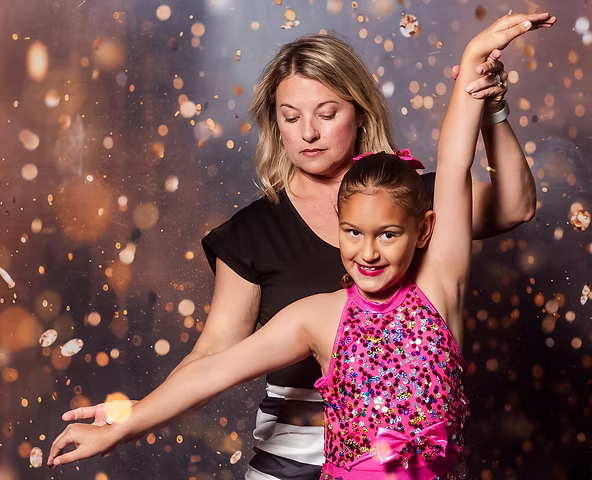 A young dancer in a sparkly pink costume practices with an instructor, who gently guides her arm placement against a warm, glowing backdrop.