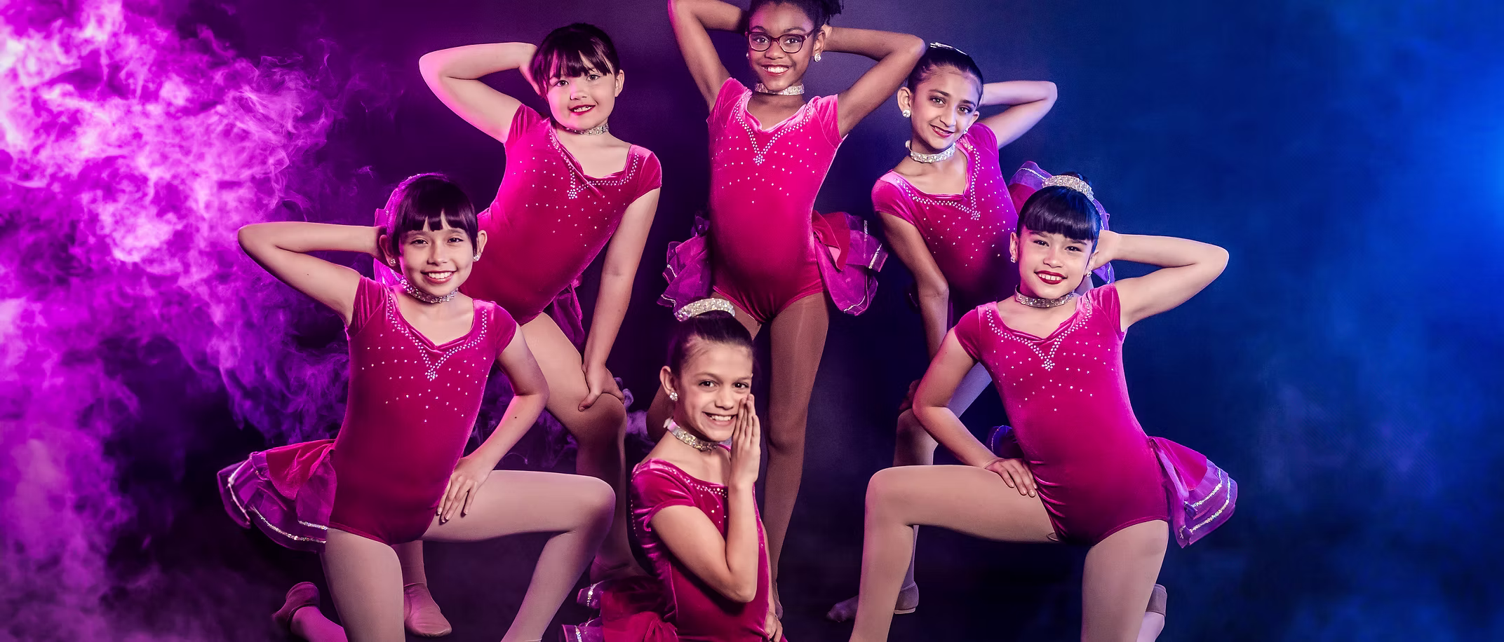 Six young dancers in bright pink costumes strike confident, playful poses onstage, surrounded by vibrant purple and blue stage lighting and theatrical smoke effects.