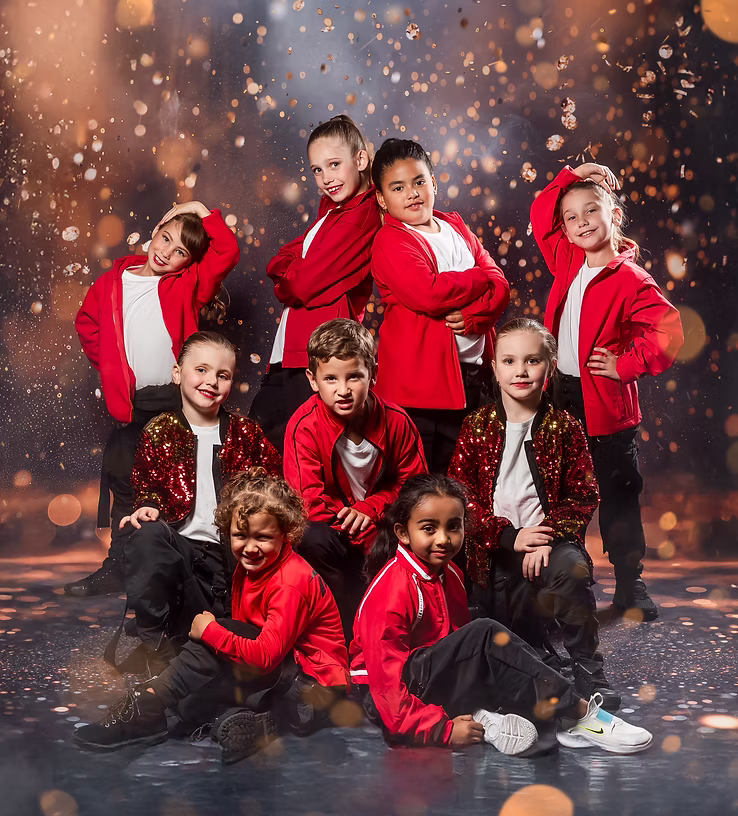 Nine dancers in red jackets and black pants pose confidently onstage, combining standing and seated levels as warm sparkles and lighting enhance their bold, energetic hip-hop vibe.