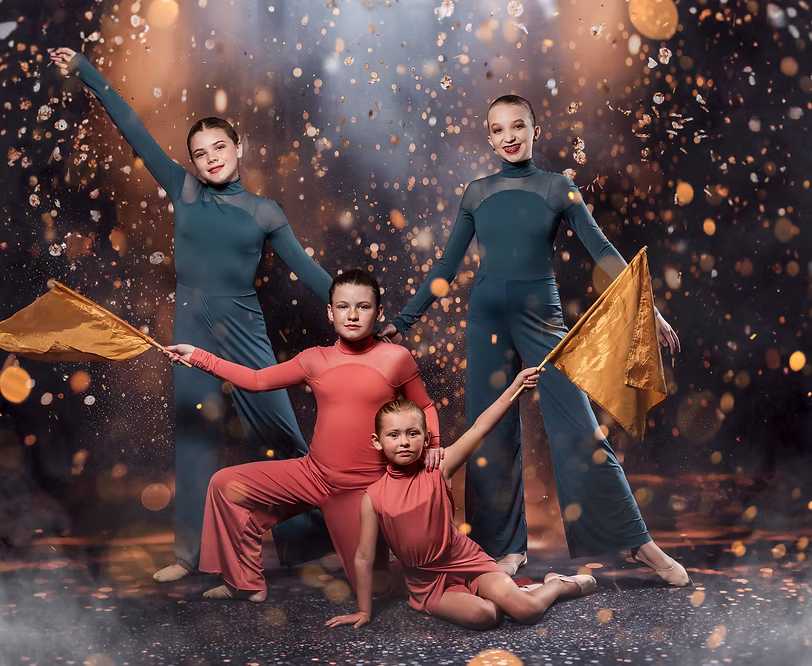 Four dancers in teal and coral costumes pose with flowing gold flags, arranged in dynamic levels as warm sparkles create a dramatic contemporary performance atmosphere.