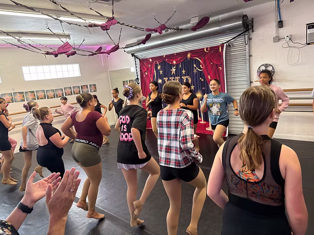 A group of teens practices dance combinations in a studio decorated with a red “VIP” backdrop, moving together in a lively, upbeat class.