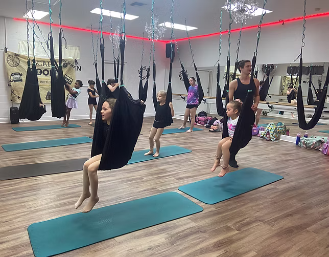 Children participate in an aerial hammock class, sitting and swinging in fabric slings above yoga mats while instructors guide them in a bright studio.