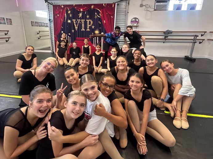 A large group of dancers in black class attire gathers for a joyful group photo, smiling and posing together in front of a “VIP” stage backdrop.