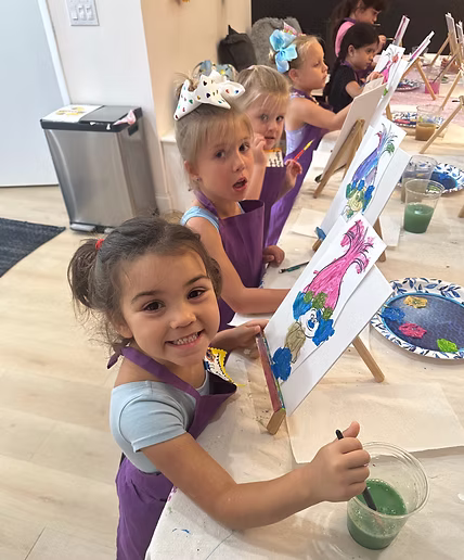 Young children wearing aprons sit at easels in an art activity, smiling and painting colorful pictures during a creative aftercare session.