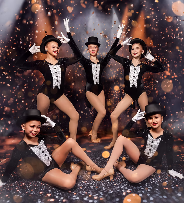 Five dancers in elegant navy dresses perform a soft, lyrical pose onstage, extending their arms gracefully as warm golden lights and sparkles create a dreamy, expressive atmosphere.