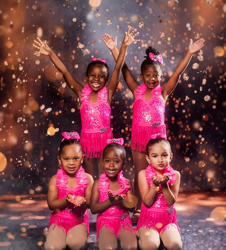 Five young dancers in bright pink sparkly costumes pose joyfully onstage, with three standing and three kneeling as warm lights and glittering effects create a cheerful, energetic atmosphere.