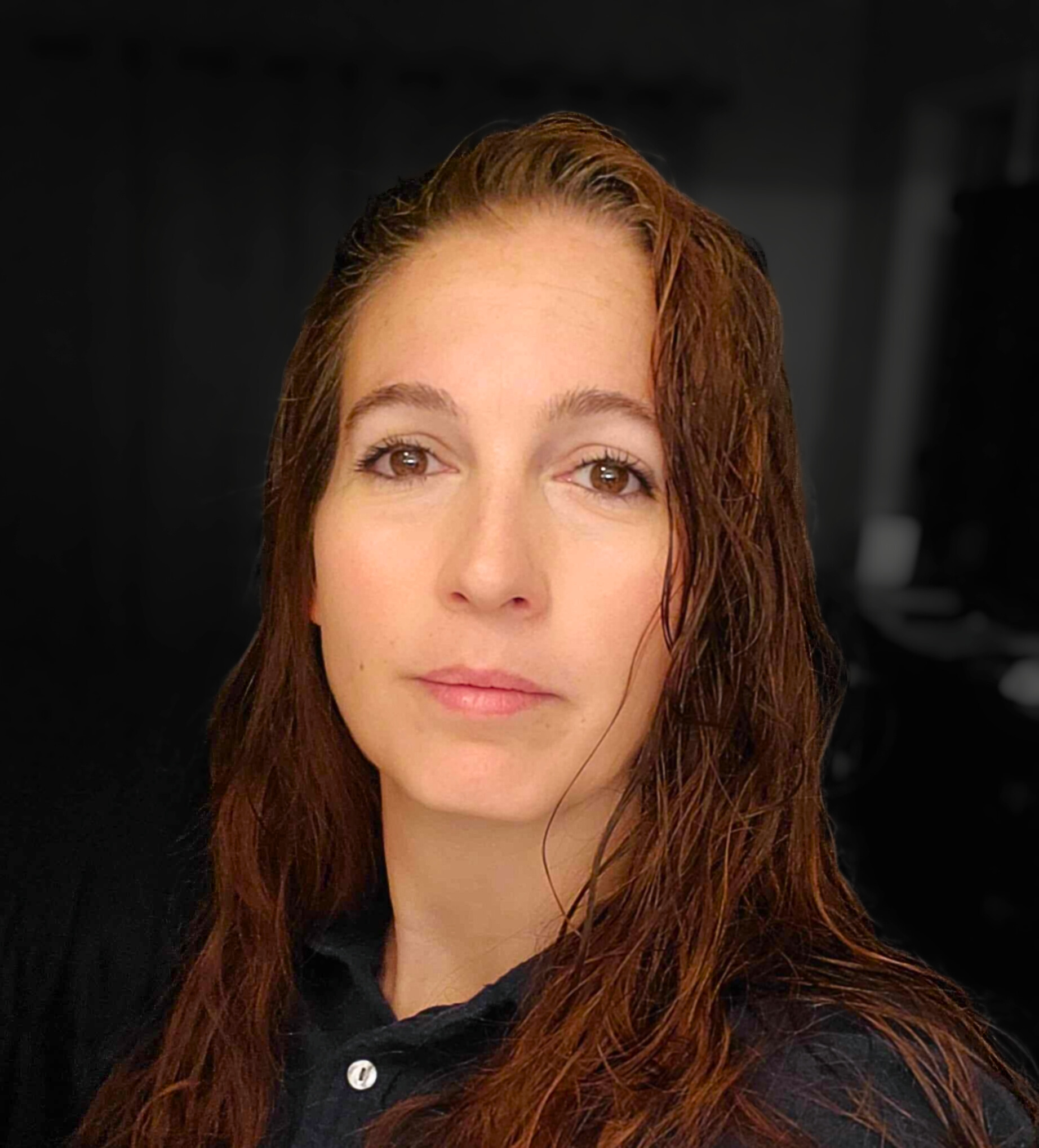 A woman with long wavy brown hair looks directly at the camera, wearing a dark top in a softly lit indoor setting, creating a natural and professional portrait