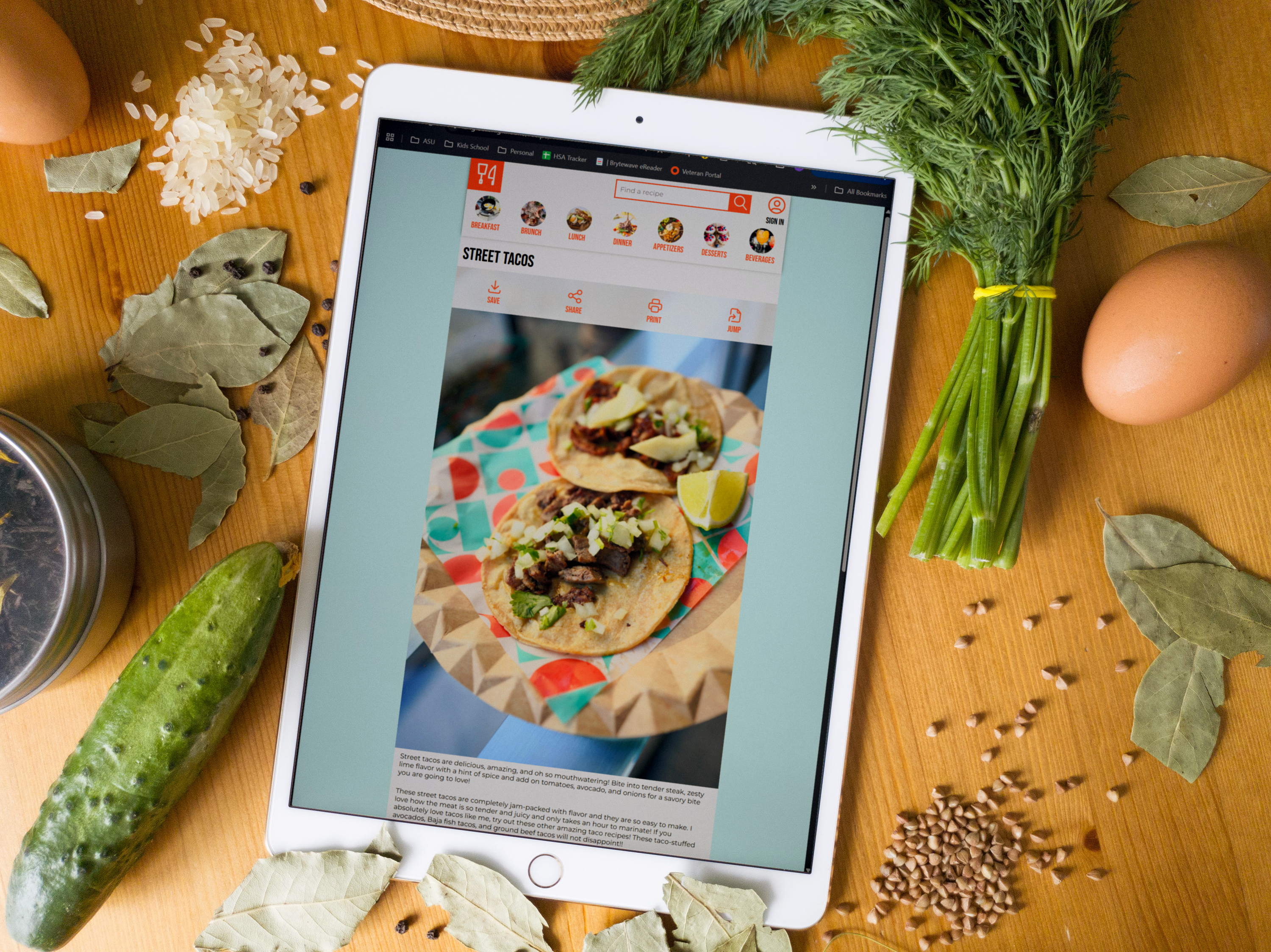 An iPad on a wooden table displays a street taco recipe, surrounded by ingredients like herbs, bay leaves, rice, cucumber, and an egg, creating a cooking-inspired presentation.