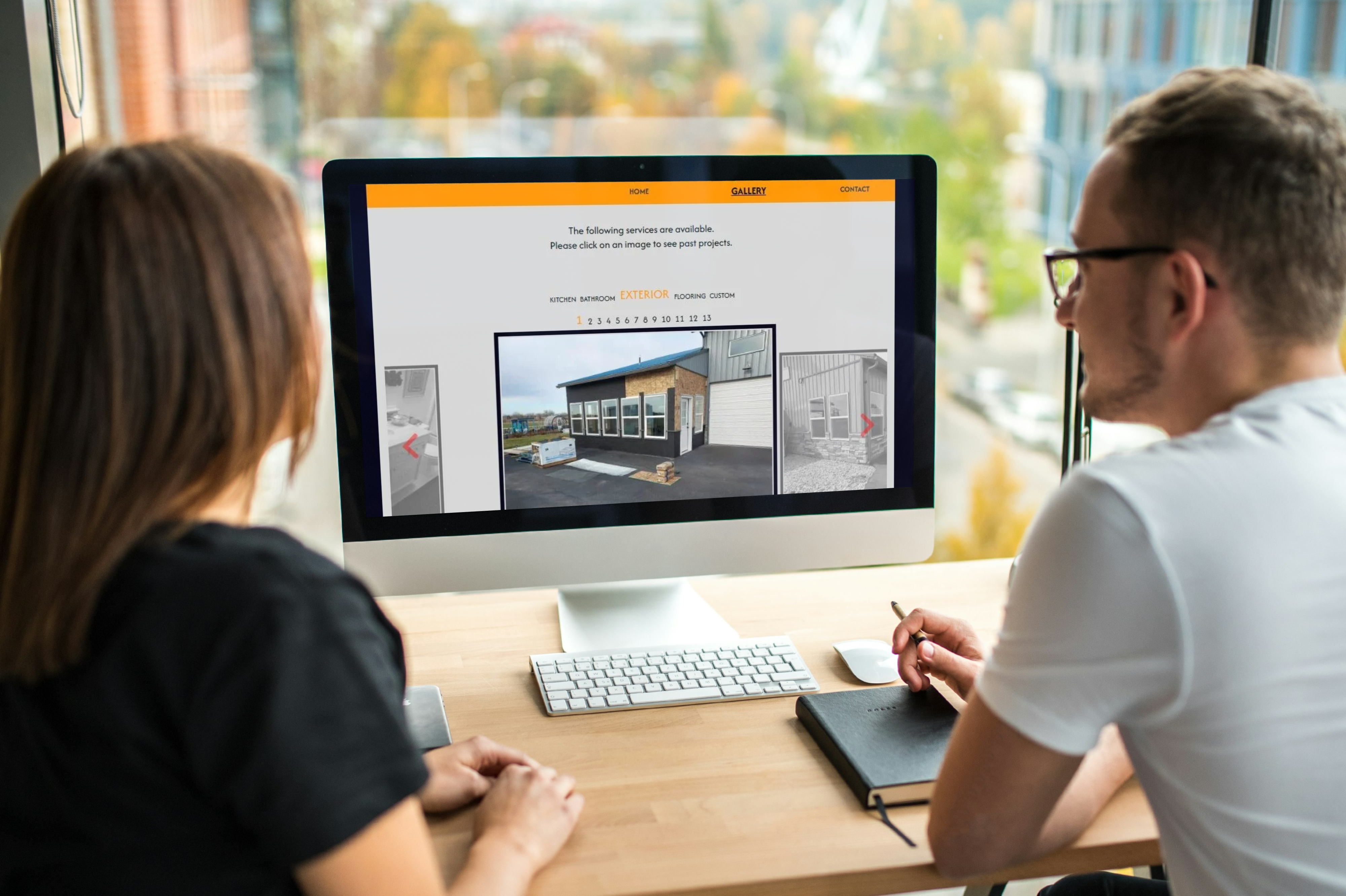Two people sit at a desk reviewing the SMC Construction website on an iMac, viewing the project gallery page and discussing exterior renovation images displayed on the screen.
