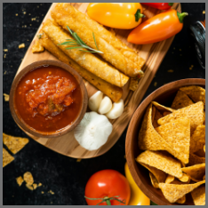 A bowl of salsa on a wooden serving tray with eggrolls, garlic and onion
